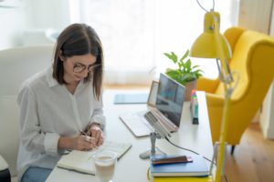 A woman writes notes in her desk, next to her computer and microphone
